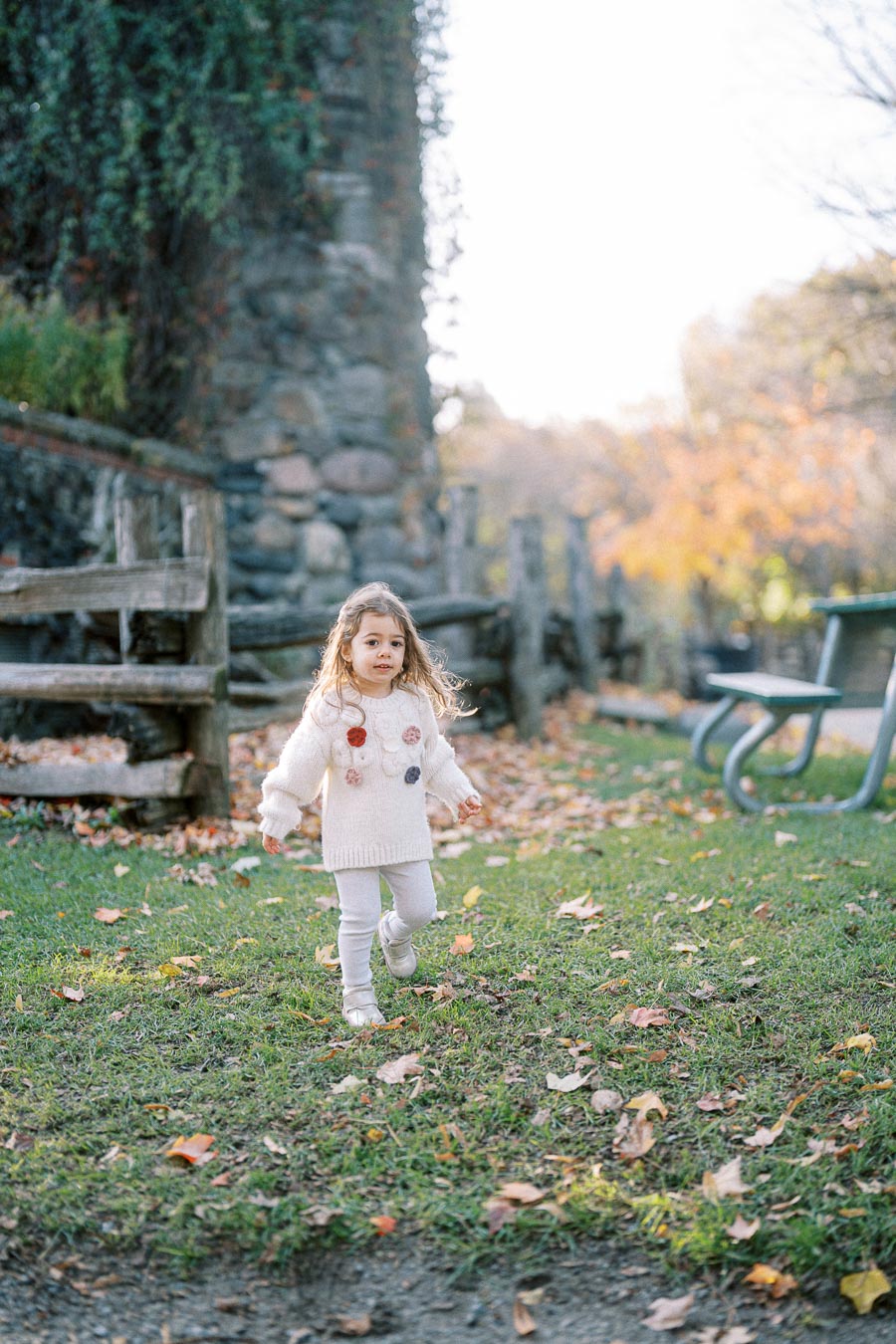 A young girl in a white sweater with colorful pom-poms runs across a grassy park area, surrounded by autumn leaves and a rustic wooden fence, with soft sunlight filtering through the trees.