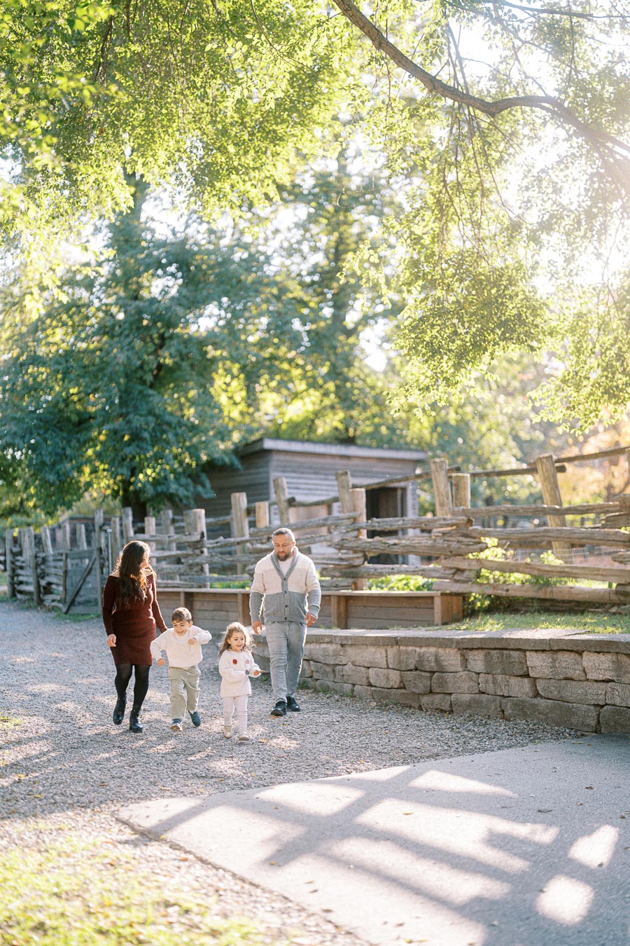 A family of four walking along a sunlit path in a picturesque park, surrounded by lush greenery and rustic wooden fencing.