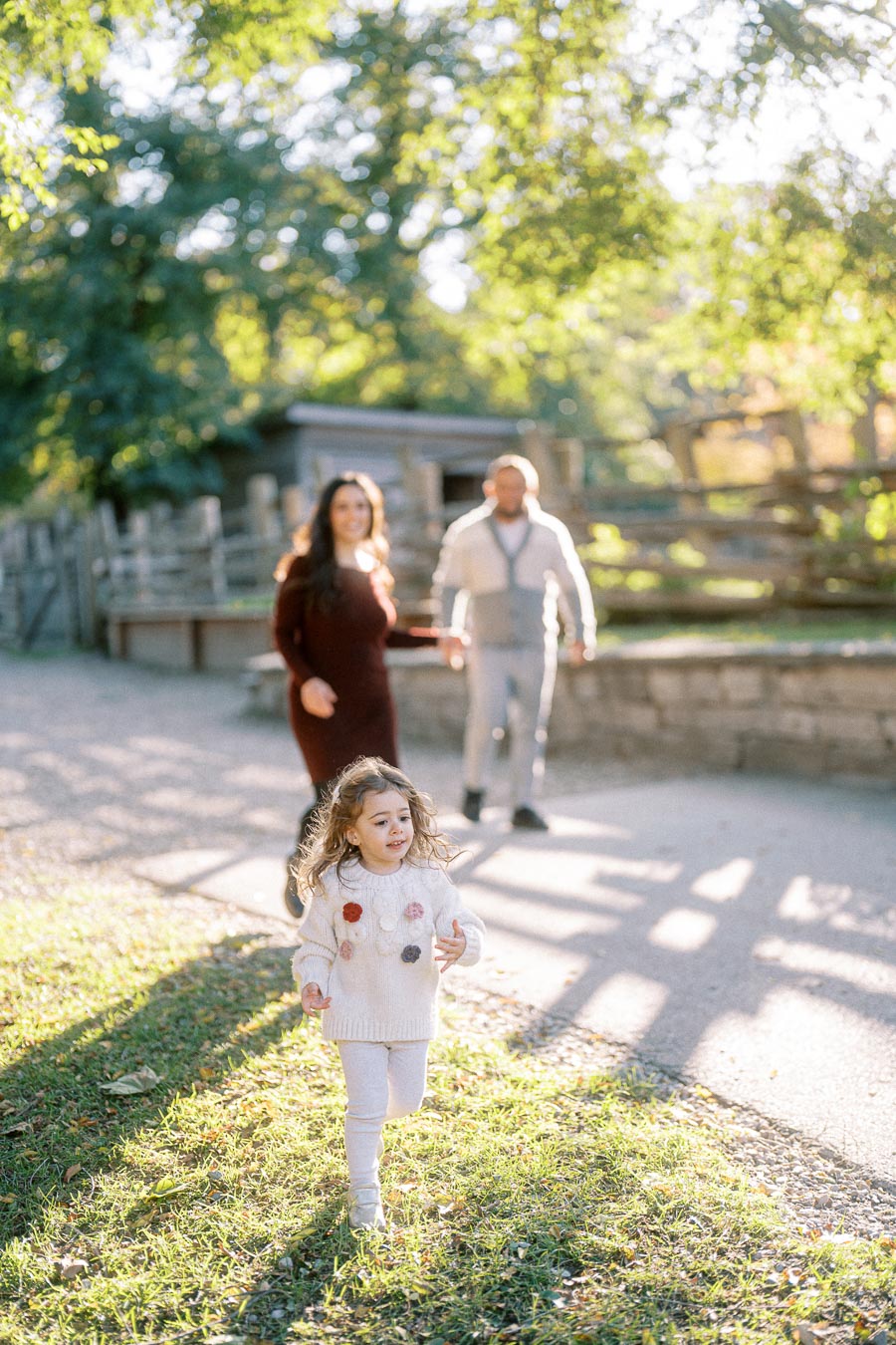 A cheerful young girl in a white sweater running on a sunlit path with her parents walking behind her, surrounded by lush greenery and a rustic wooden fence.