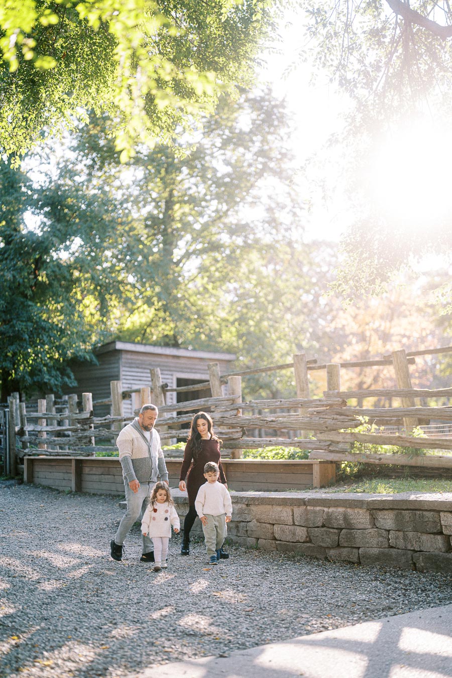 A family walking together on a sunny day in a rustic park setting, with wooden fences and lush green trees in the background.