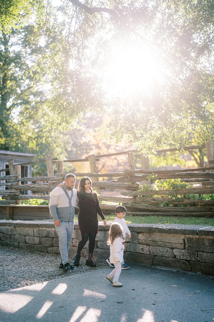 A family of four enjoys a sunny day outdoors, walking along a rustic wooden fence lined pathway.