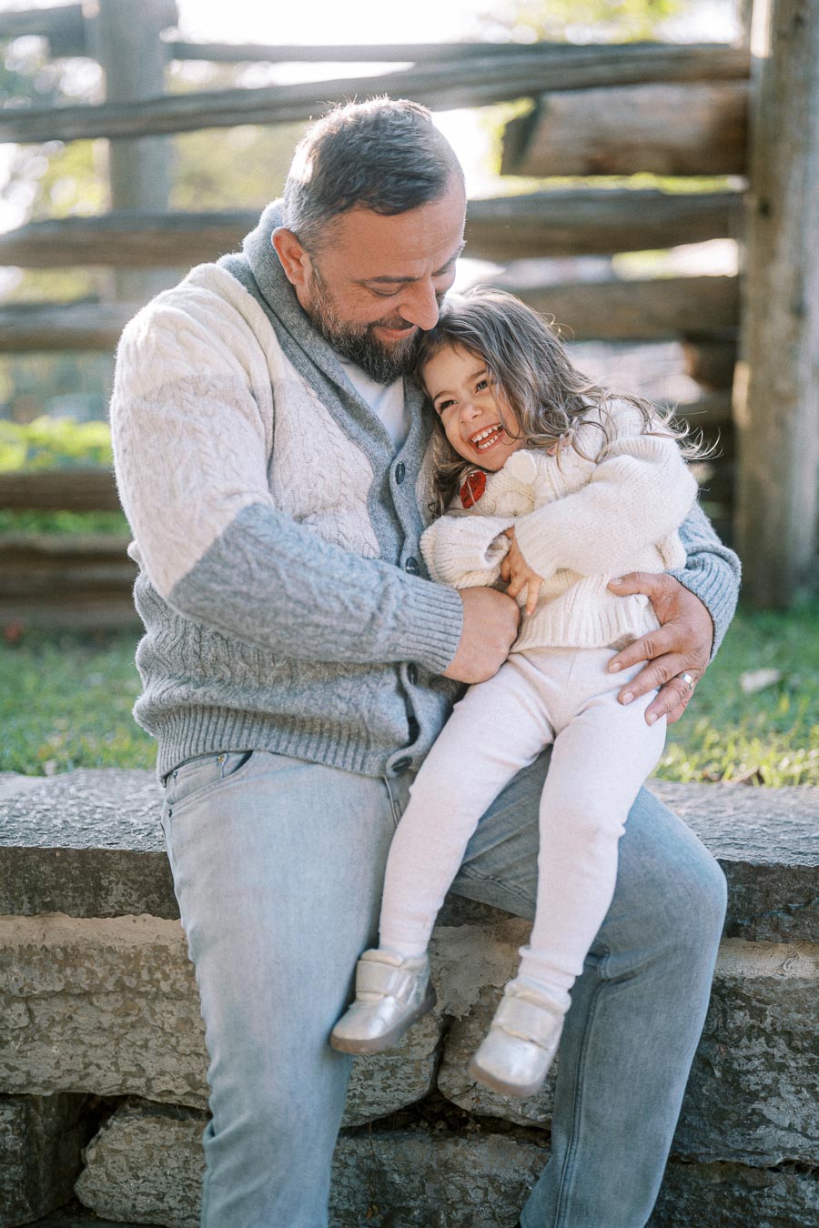 Father and daughter sharing a joyful moment outdoors, sitting on a stone bench in warm knit sweaters.