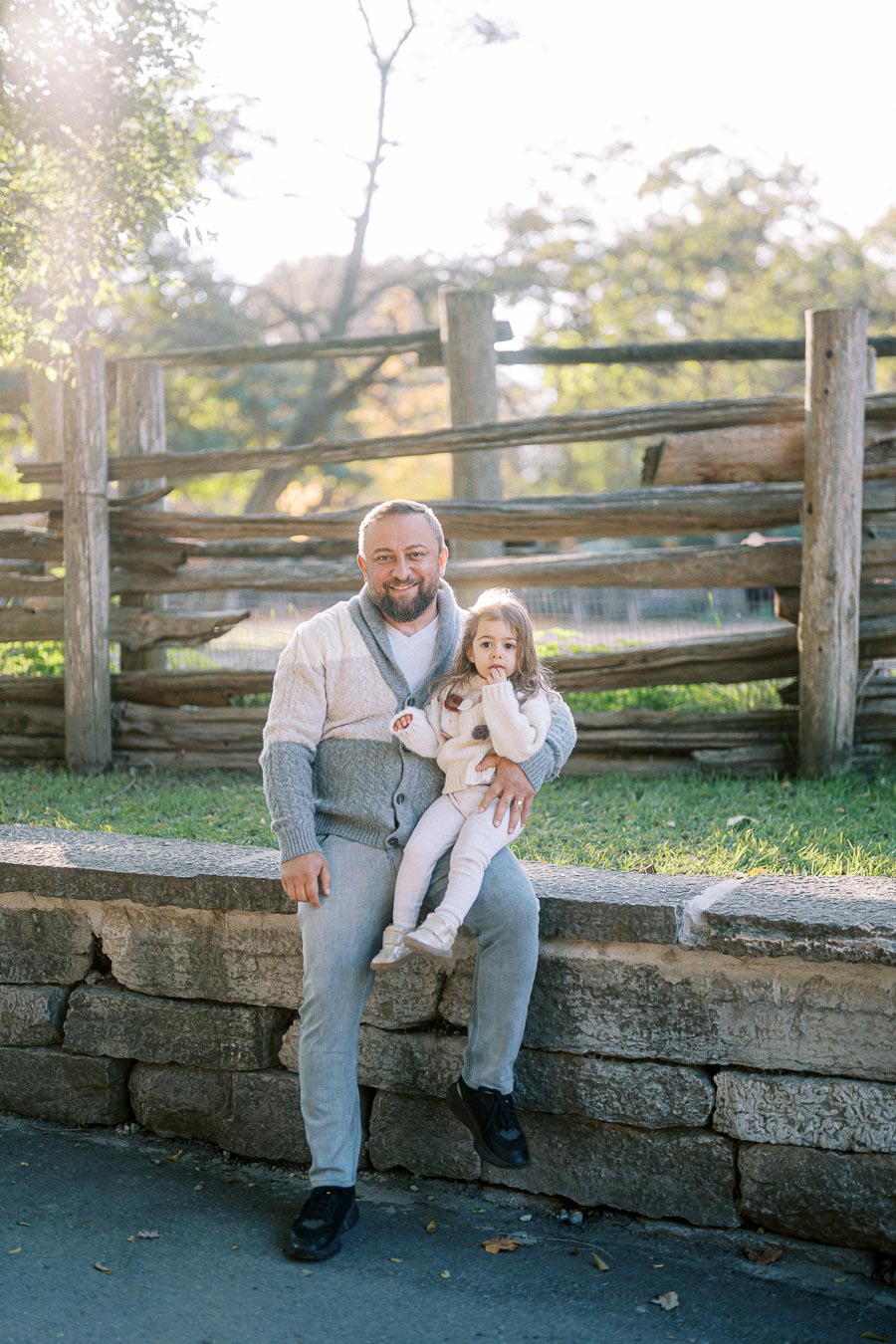 A smiling man sitting on a stone wall with a young girl on his lap, surrounded by a rustic wooden fence and greenery, enjoying a sunny day outdoors.