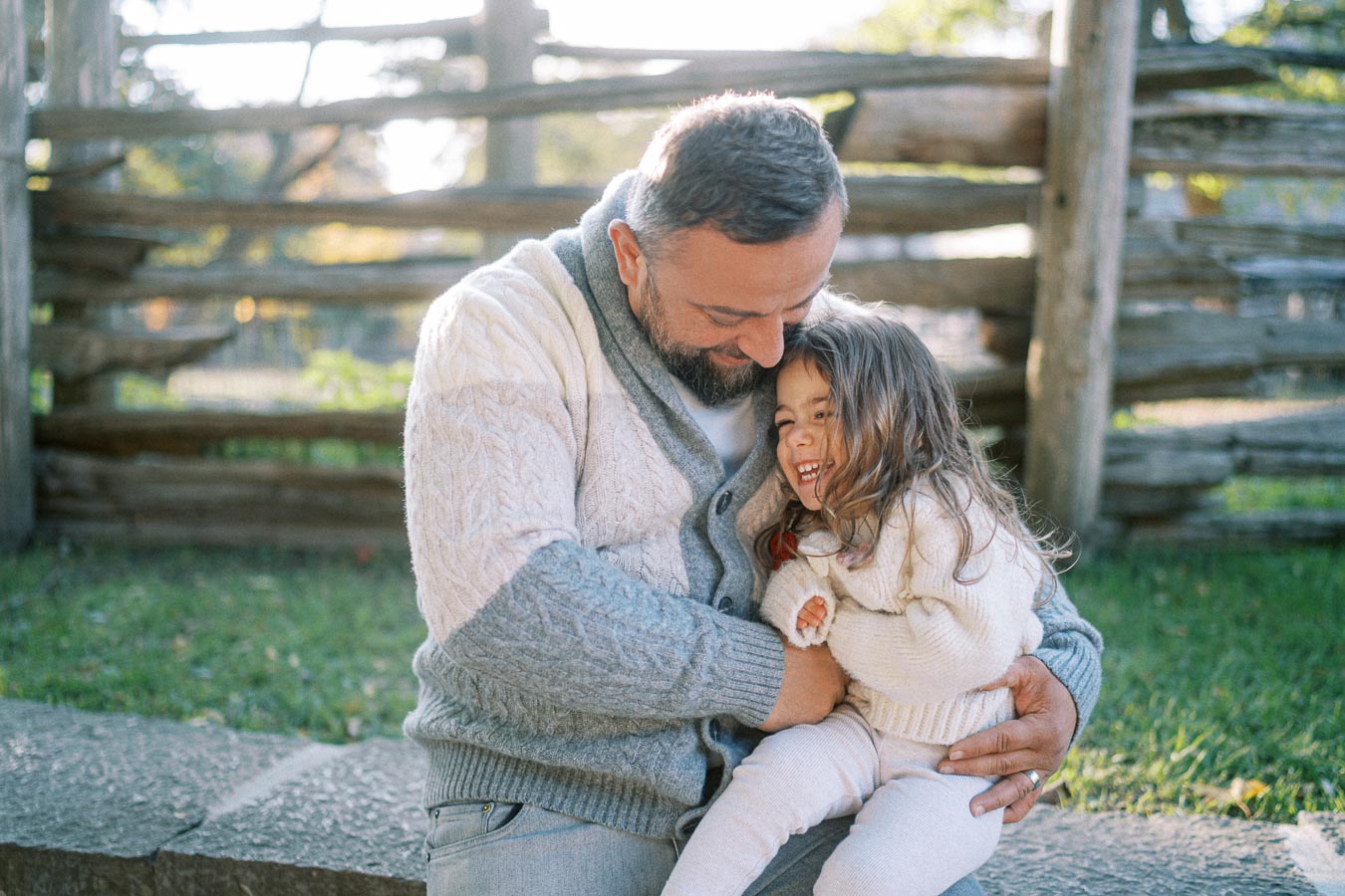 A father and daughter share a joyful moment outdoors, seated on a stone bench with wooden fencing in the background. Both are wearing cozy sweaters, enjoying a sunny day in a park.
