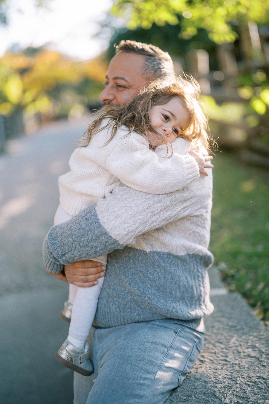 A loving father embraces his daughter in a sunny park, both in cozy sweaters, with autumn foliage in the background.
