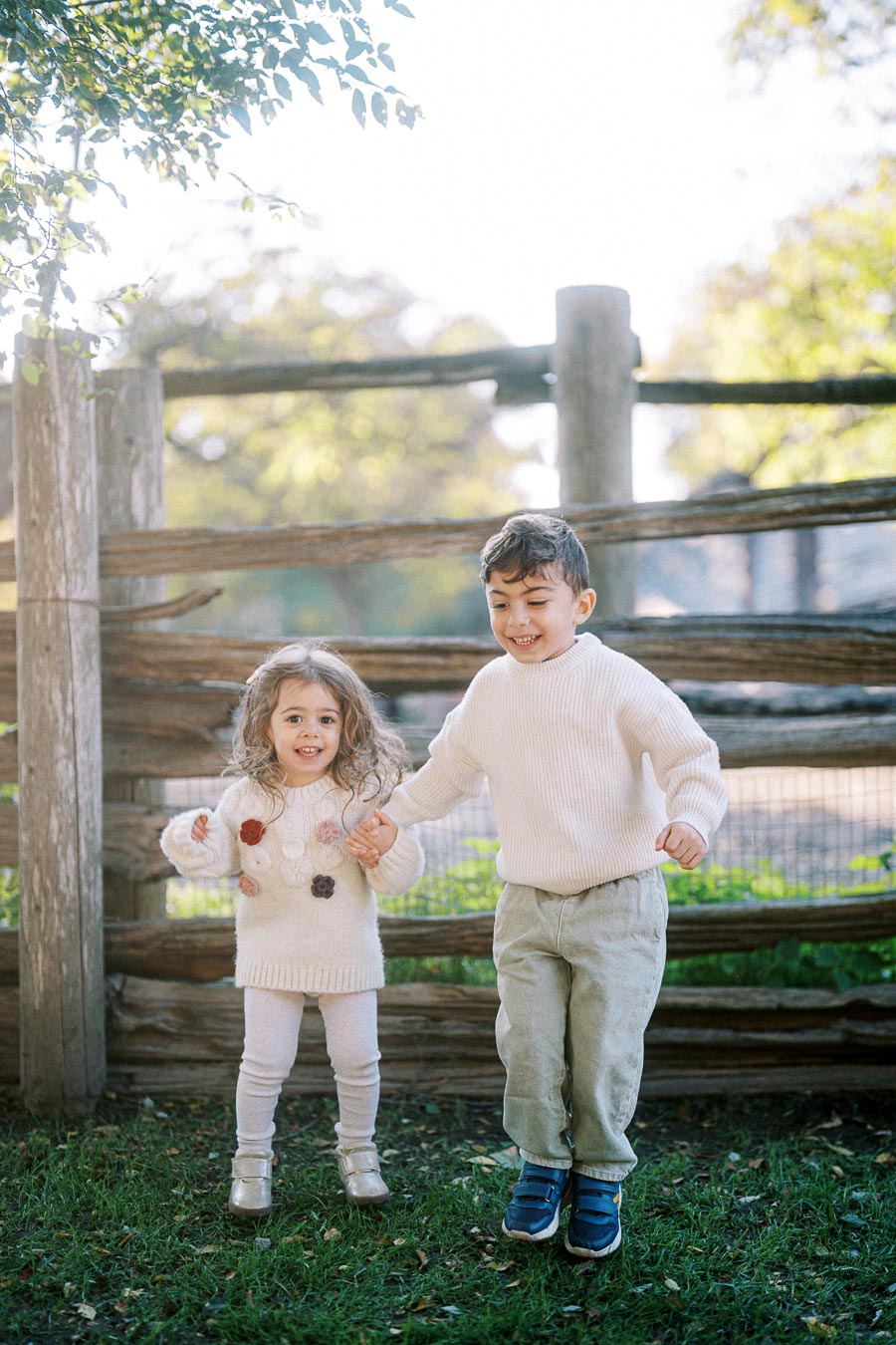Two young children, a boy and a girl, joyfully jumping in front of a wooden fence in a sunlit park. The children are wearing cozy sweaters and appear to be enjoying a bright, clear day surrounded by lush greenery.