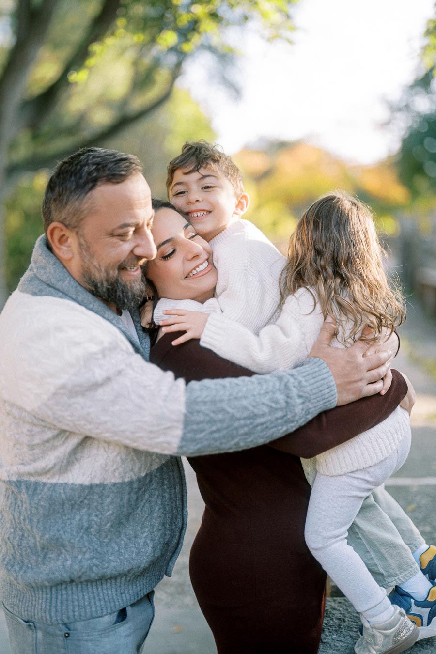 A happy family of four sharing a warm embrace outdoors on a sunny day, surrounded by lush green trees, wearing cozy sweaters and smiling brightly.