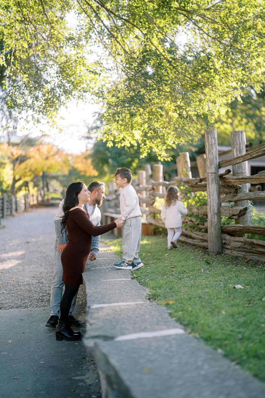 Family enjoying a sunny day in the park, with parents interacting with their children near a wooden fence surrounded by greenery.