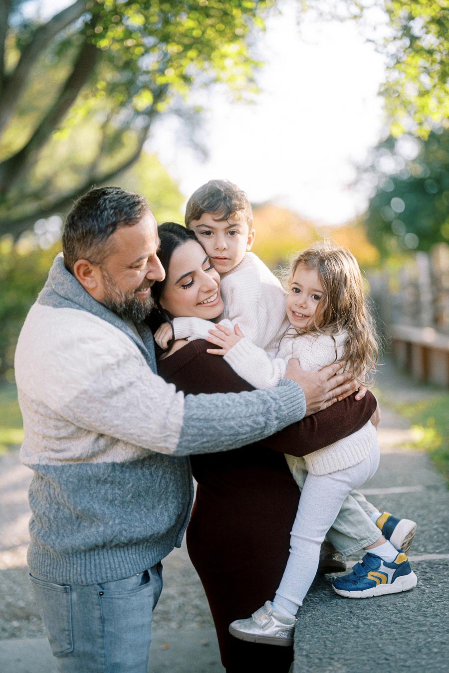 A happy family of four embracing in a sunny park, with green foliage in the background, wearing cozy sweaters and smiling together.