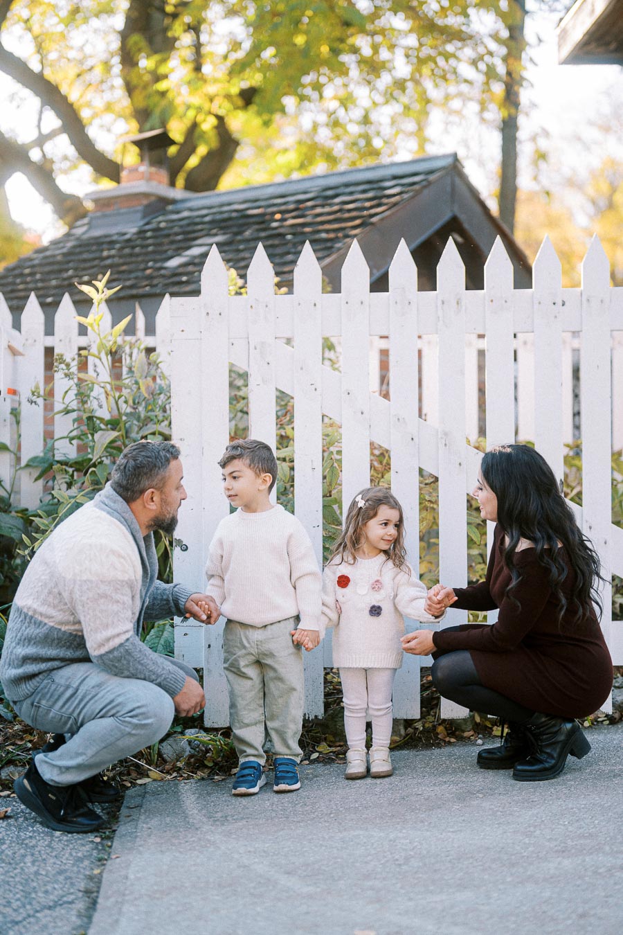 Family enjoying a sunny day outdoors by a white picket fence, with a father holding his son's hand and a mother holding her daughter's hand, surrounded by autumn foliage.