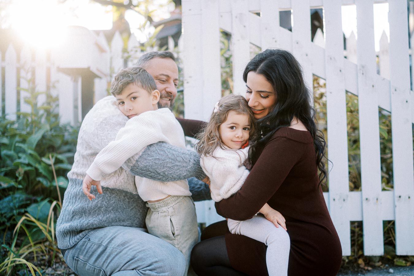 A happy family with two children hugging outdoors in front of a white picket fence, with sunlight filtering through trees in the background.