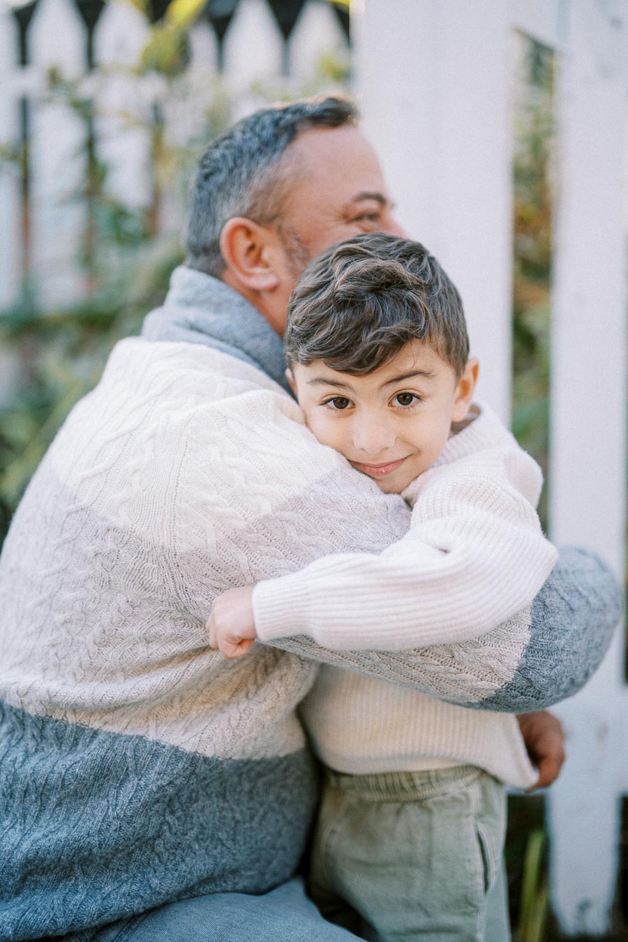 Father and son embracing warmly outdoors, both wearing cozy sweaters, with a white picket fence in the background.