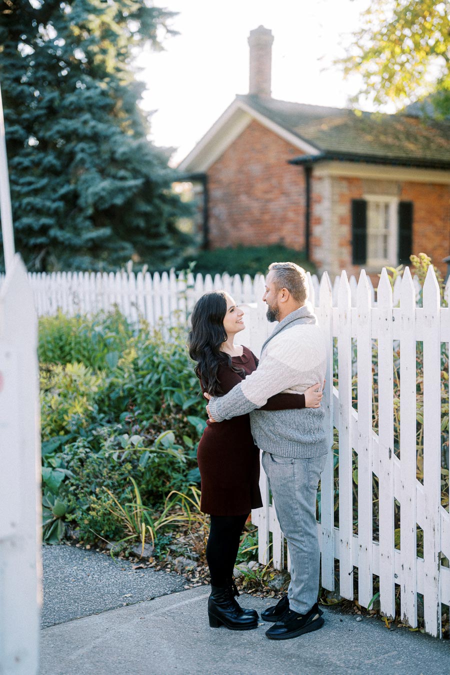 Couple embracing near a white picket fence with a brick house and lush greenery in the background, showcasing a warm and romantic outdoor scene.
