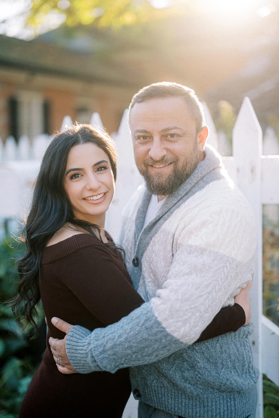 Happy couple embracing near a white picket fence, backlit by the sun, enjoying a sunny day outdoors.