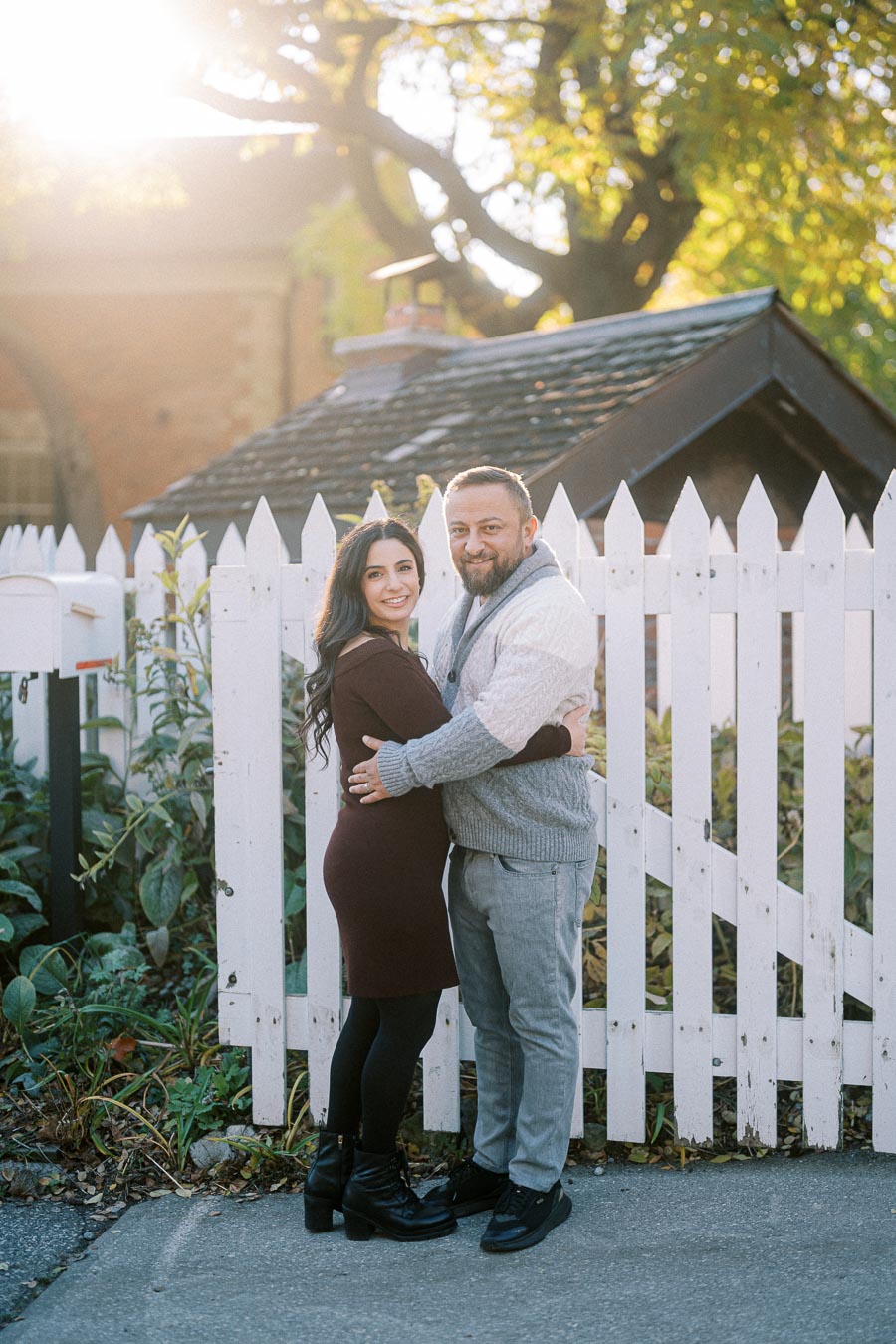 A couple embracing in front of a white picket fence on a sunny autumn day, with sunlight filtering through the trees in the background.
