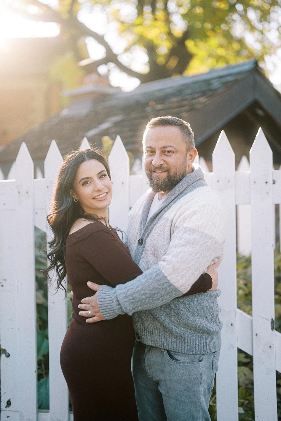A smiling couple embracing in front of a white picket fence, bathed in warm, natural sunlight, with green foliage and a quaint wooden structure in the background.