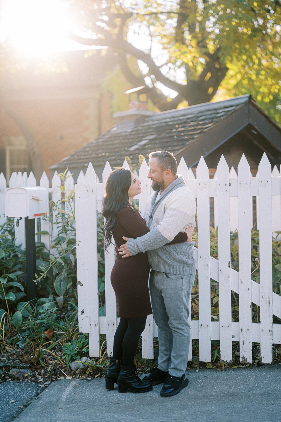 A couple embraces lovingly in front of a white picket fence with autumn foliage and sunlight streaming through the trees, conveying warmth and romance in an outdoor setting.