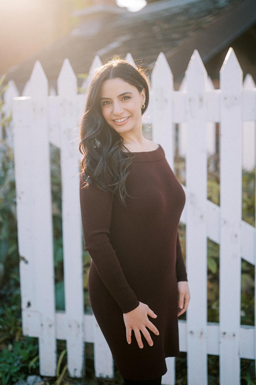 Smiling woman in a burgundy dress standing in front of a white picket fence with sunlight in the background.