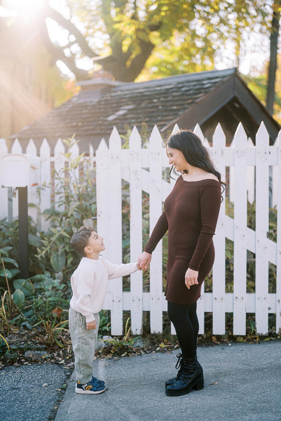 Mother and son holding hands in front of a white picket fence in an autumn setting. The mother is wearing a brown dress and black boots, while the son is in a white sweater and light pants. Bright sunlight filters through the trees, creating a warm, inviting atmosphere.
