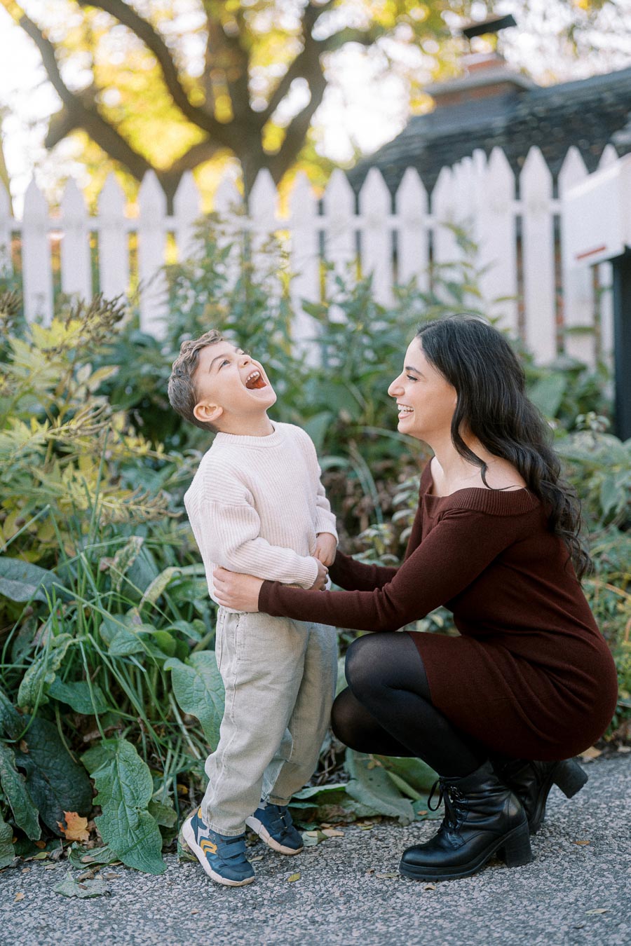 Mother kneeling and laughing with her young son in a garden, surrounded by greenery and a white picket fence.