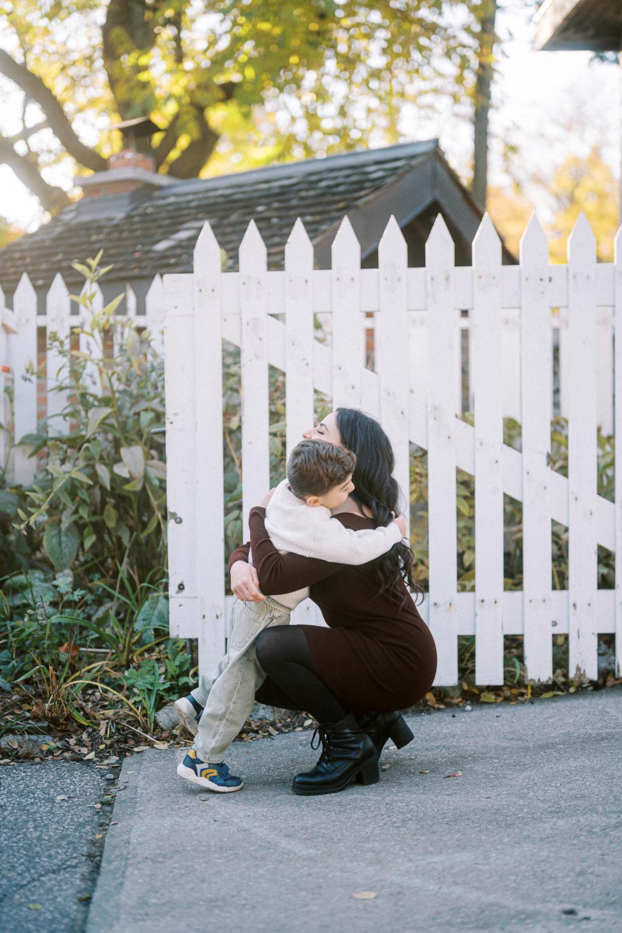 A woman crouches to embrace a young boy in front of a white picket fence, surrounded by greenery and illuminated by soft daylight.