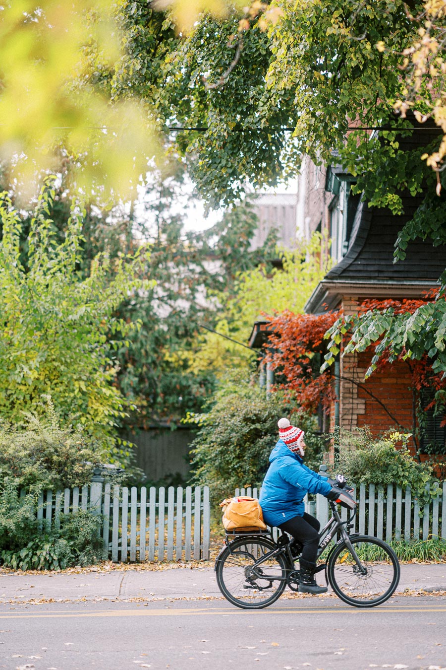 A person in a blue jacket and red-and-white knit hat rides a bicycle along a tree-lined street with a white picket fence and autumn foliage in the background.