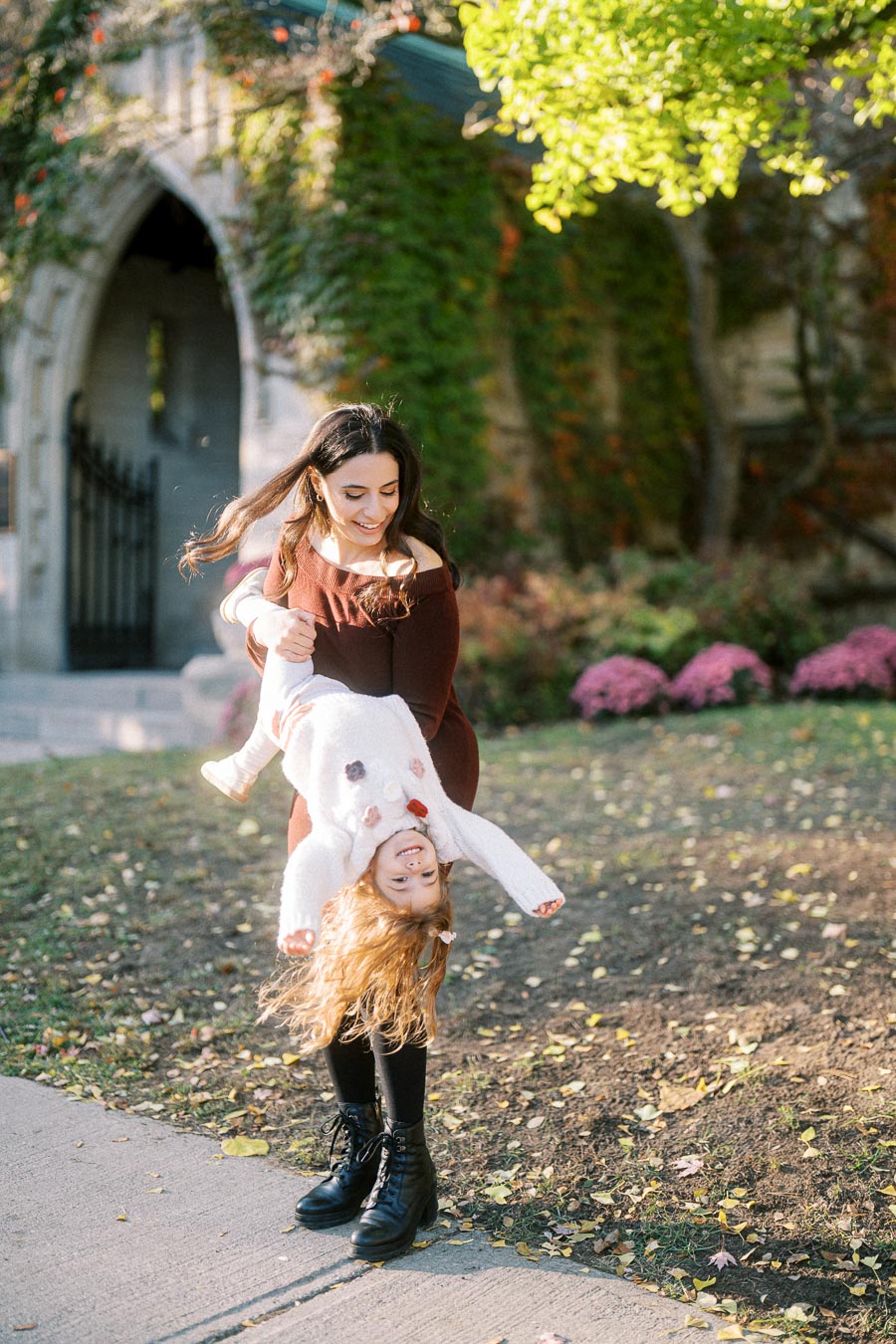 A woman playfully swings a young girl upside down in a park setting with greenery and a stone building in the background. The scene captures a joyful moment amidst colorful autumn foliage.