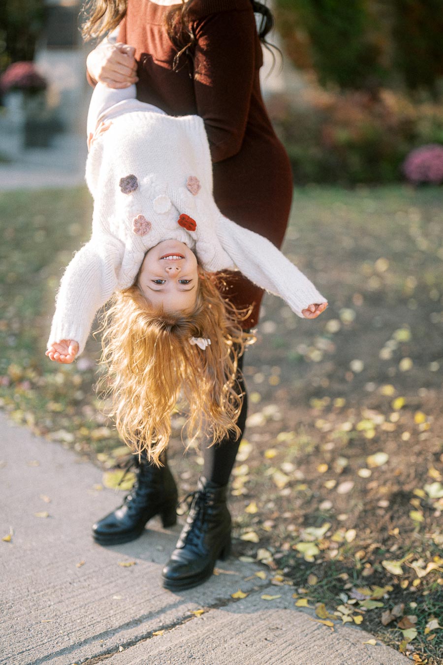 A young child with curly blonde hair playfully hangs upside down, held by an adult in a brown outfit, on a sunlit sidewalk surrounded by autumn leaves.