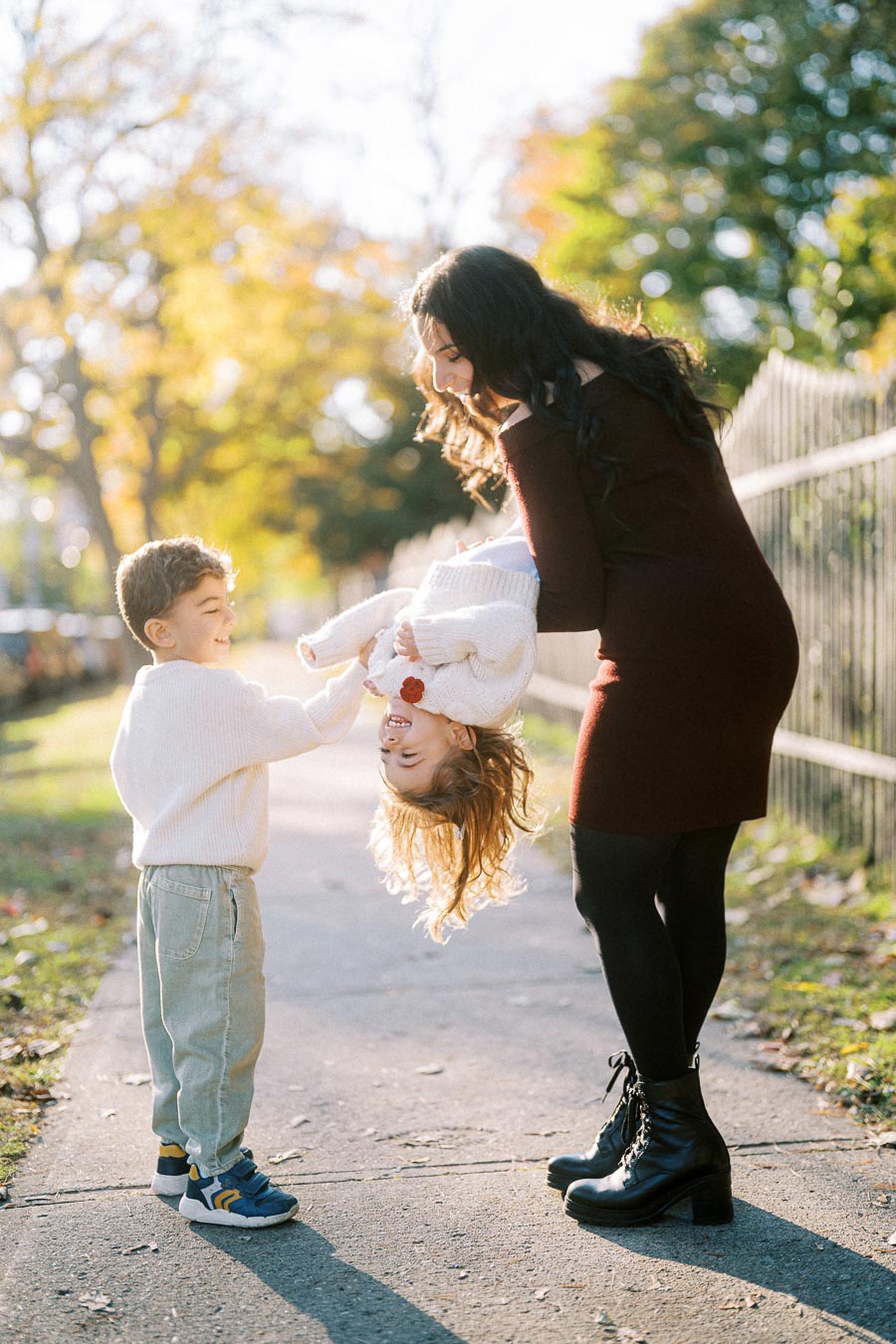A woman and two children playfully interact on a sunny sidewalk surrounded by autumn trees, with one child being gently lifted upside down, all smiling and enjoying their time outdoors.
