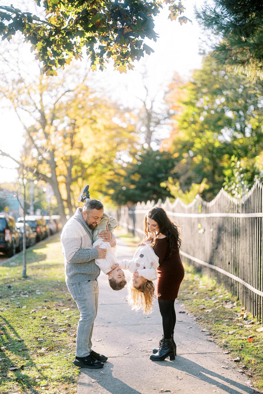A happy family enjoying a playful moment outdoors in a sunlit park, with parents holding their two young children upside down, surrounded by autumn foliage.