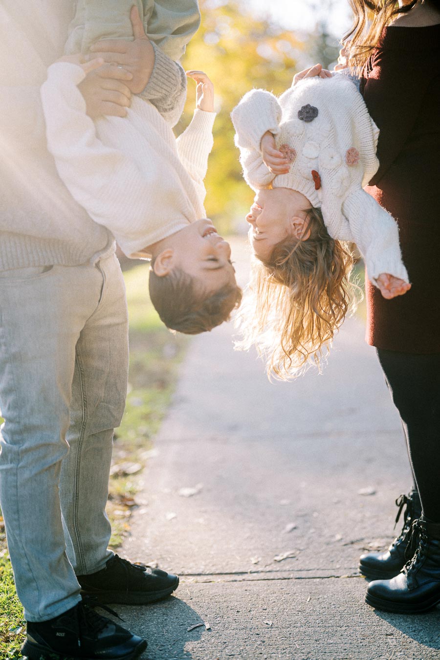 A playful autumn scene with two children joyfully hanging upside down while being held by two adults on a sunny pathway, surrounded by vibrant fall foliage.