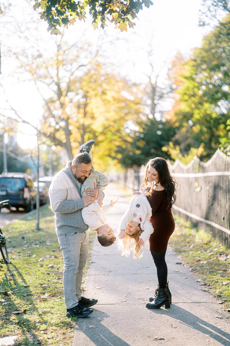 A joyful family moment in the park during autumn, featuring a father and mother playfully holding their two children upside down, with sunlight filtering through the trees.