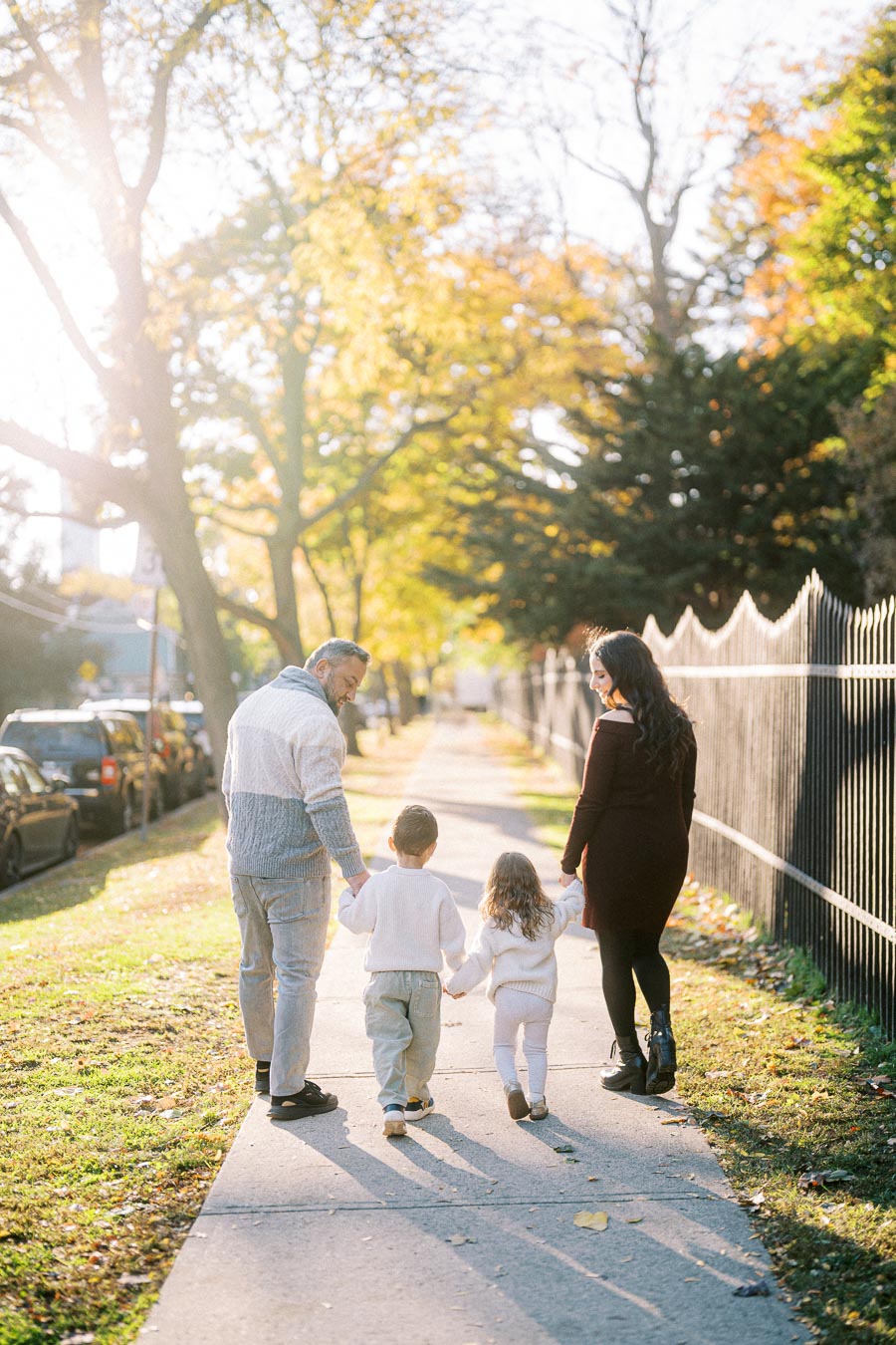 A family of four walking along a tree-lined sidewalk during autumn, with colorful leaves and sunlight filtering through the branches.