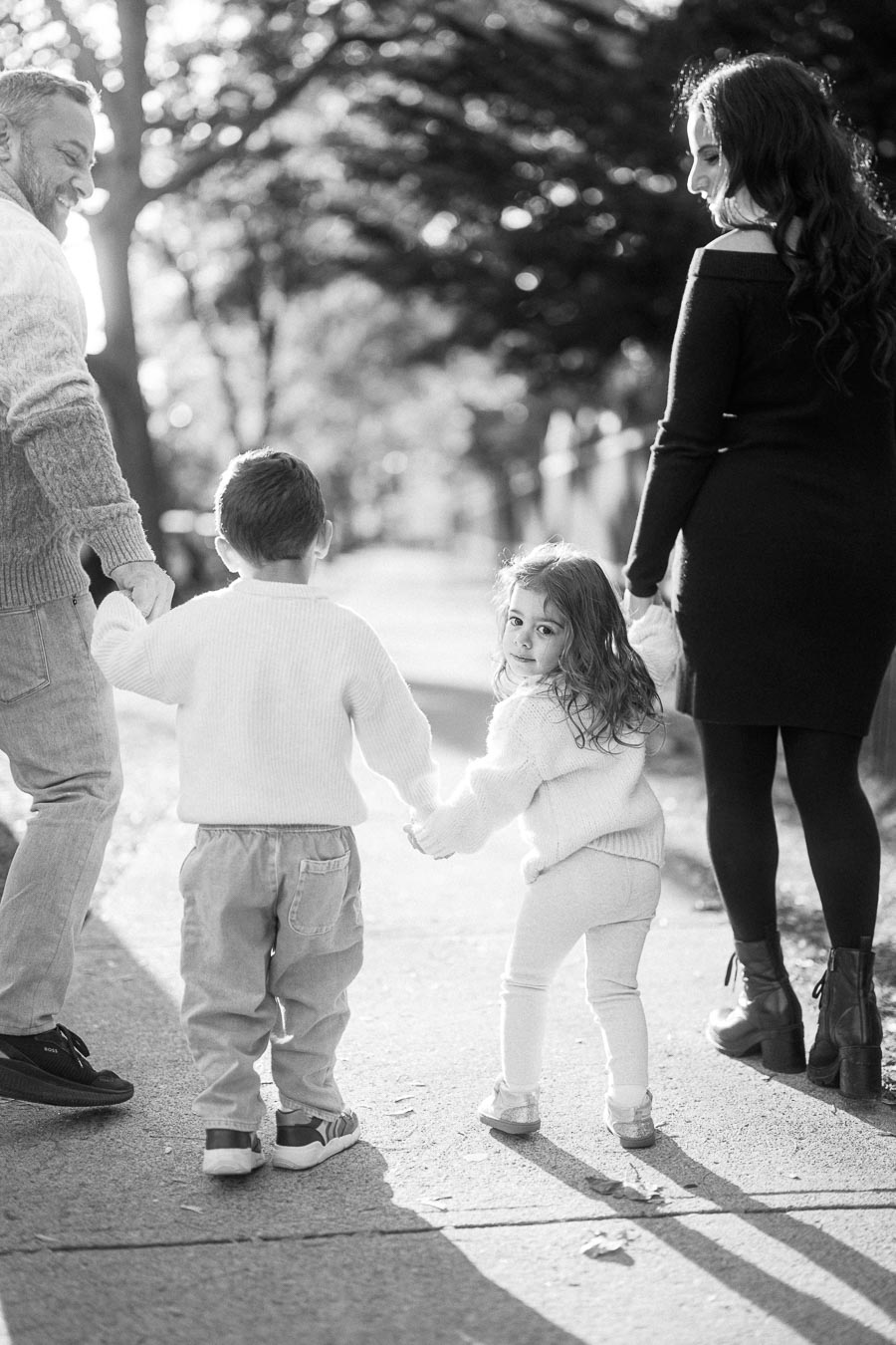 A black and white image of a family walking down a sunny sidewalk, featuring a young boy and girl holding hands with their parents as they enjoy a pleasant day out.