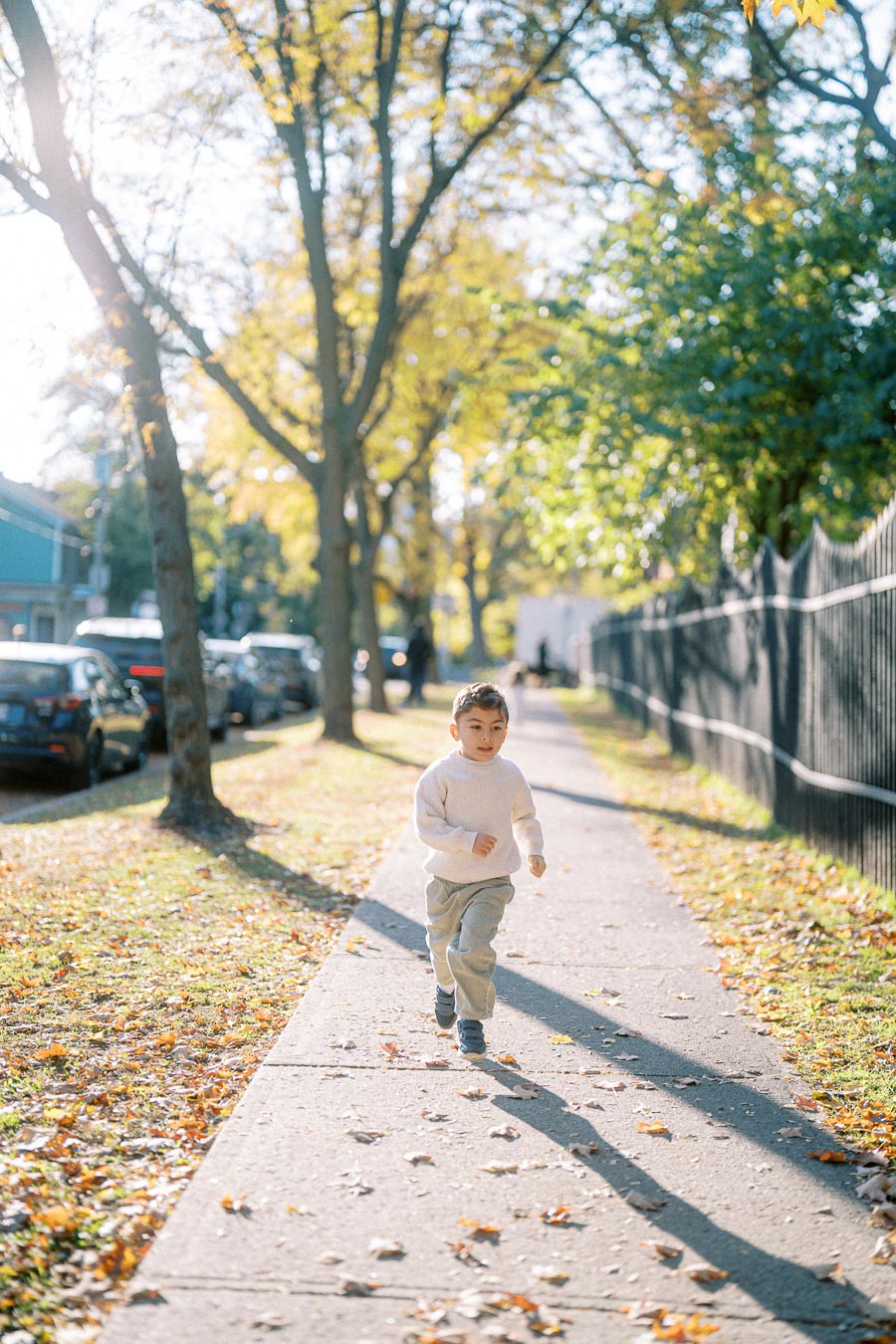 A young child in a light sweater joyfully runs along a sunlit, tree-lined sidewalk on a crisp autumn day, with scattered leaves decorating the path and parked cars in the background.