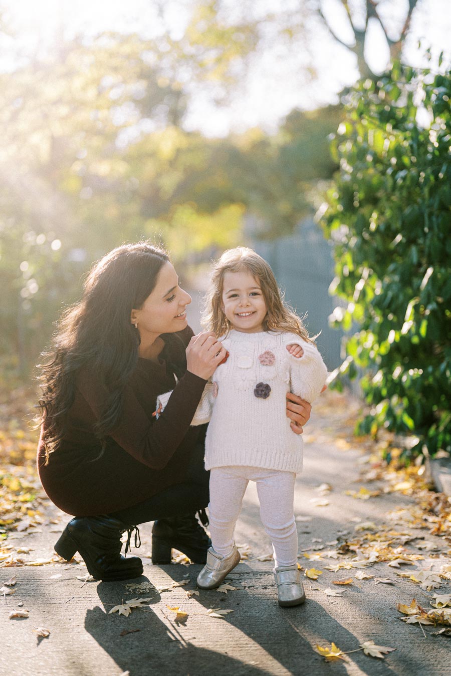A mother and daughter smiling in a sunlit park with fallen leaves and greenery, wearing cozy fall outfits.