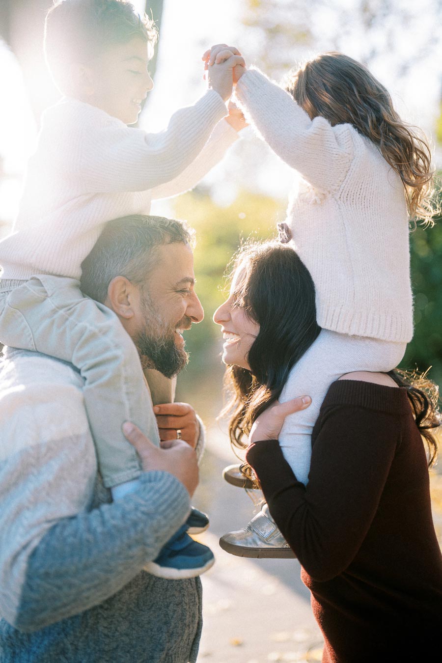 A joyful family enjoying outdoor time together, with parents carrying their smiling children on their shoulders, surrounded by warm natural lighting and green foliage.