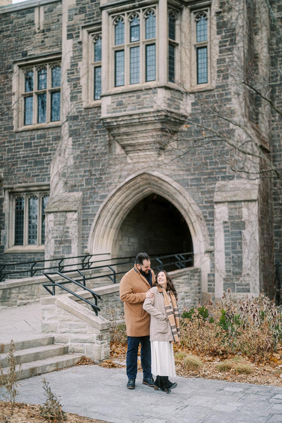 A couple stands outside a historic stone building with arched windows, dressed in warm winter coats, embracing and smiling while enjoying a romantic moment.