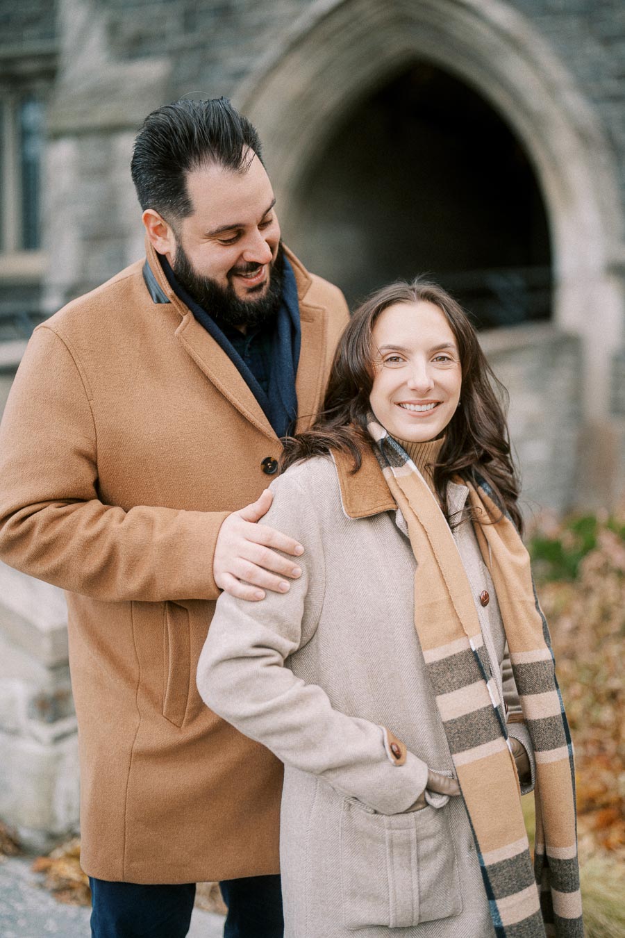 A smiling couple in winter coats enjoying a walk outdoors, with an archway and stone building in the background.