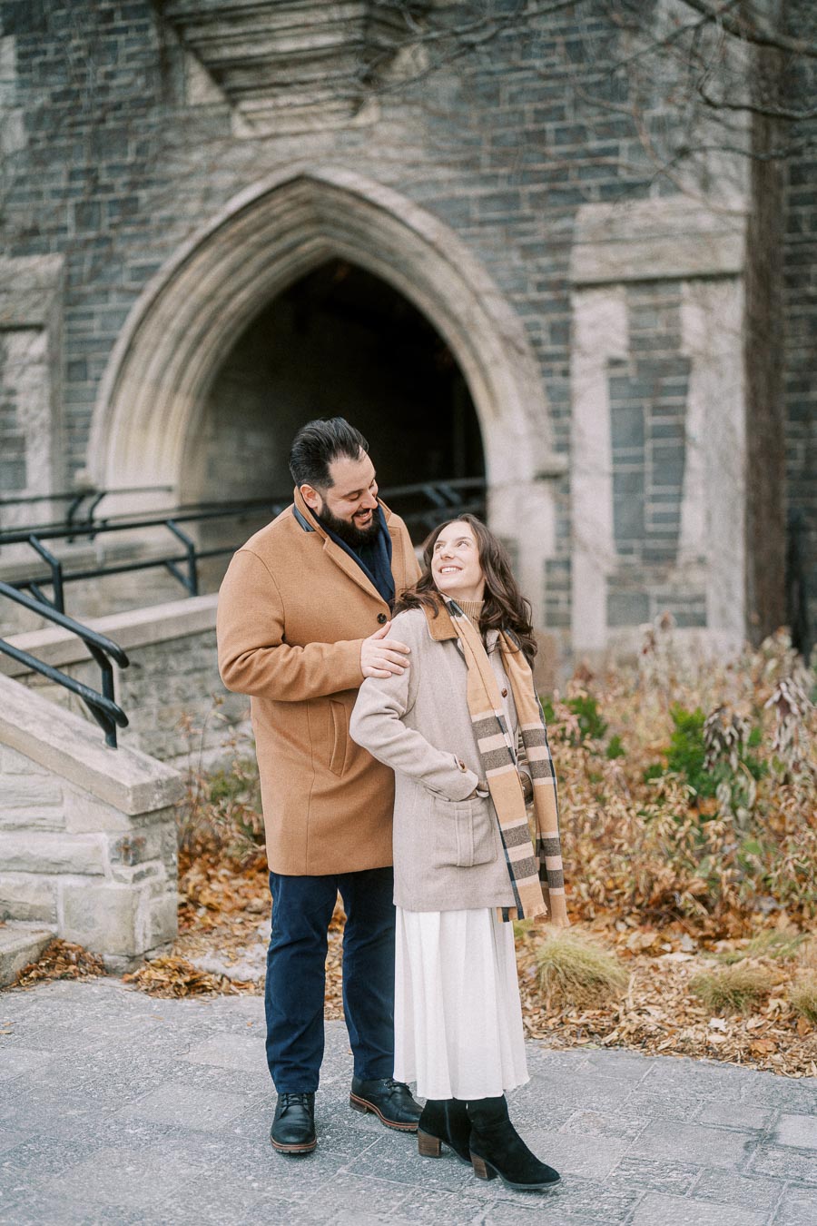 A couple in winter coats standing affectionately outside a stone building with an arched doorway, surrounded by autumn foliage.