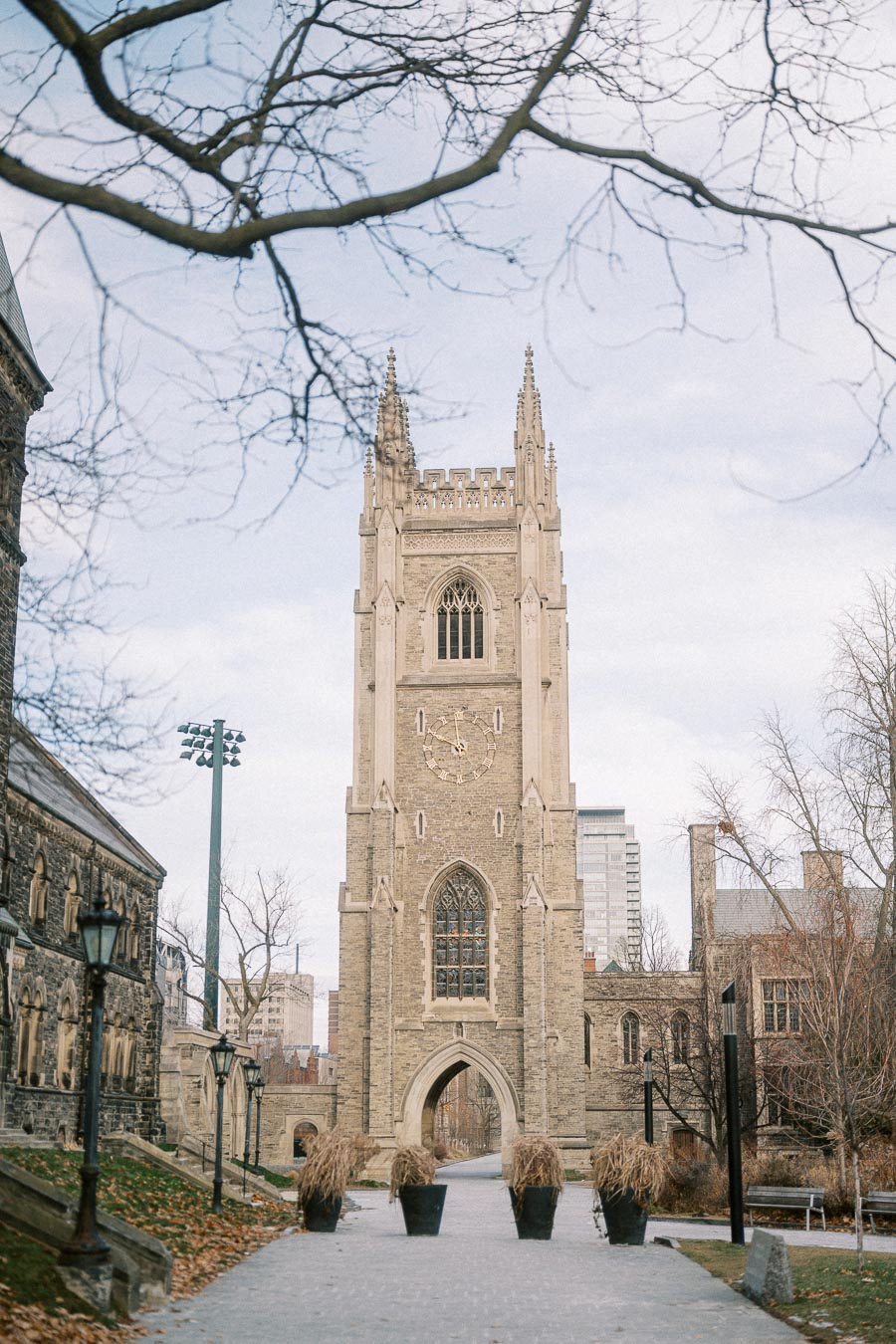 Historic stone clock tower of University College at the University of Toronto, surrounded by bare trees and vintage streetlamps on a cloudy day.