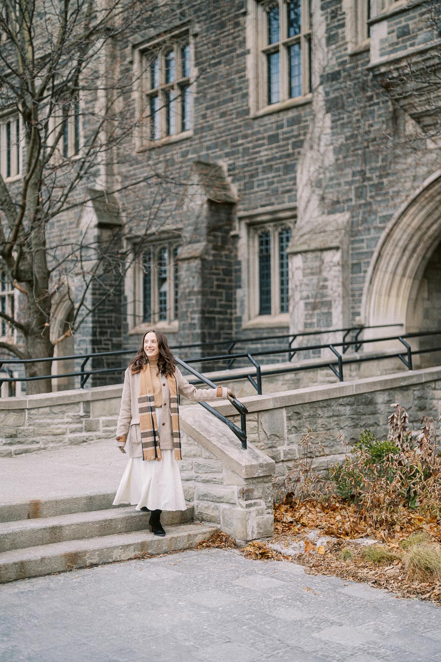 A woman in a beige coat and scarf standing on stone steps in front of a historic brick building, with fall leaves scattered on the ground.