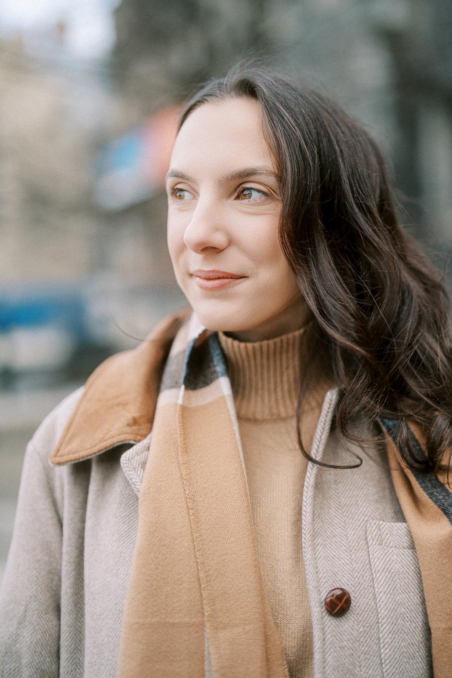 A woman wearing a beige coat and scarf smiles while looking off to the side against a blurred outdoor background.