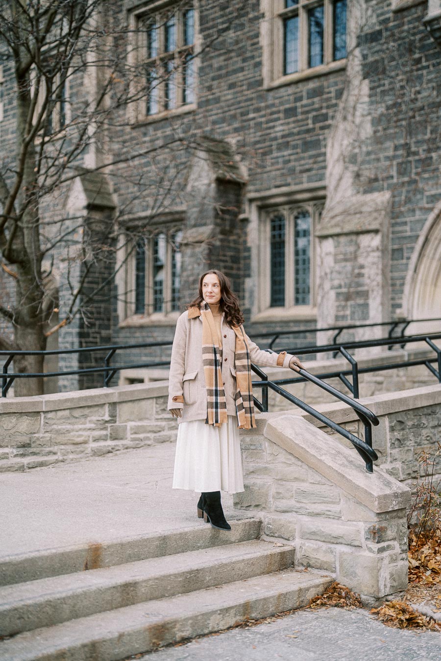 Woman in a stylish beige coat with a plaid scarf stands on stone steps near a historic brick building, showcasing autumn fashion.