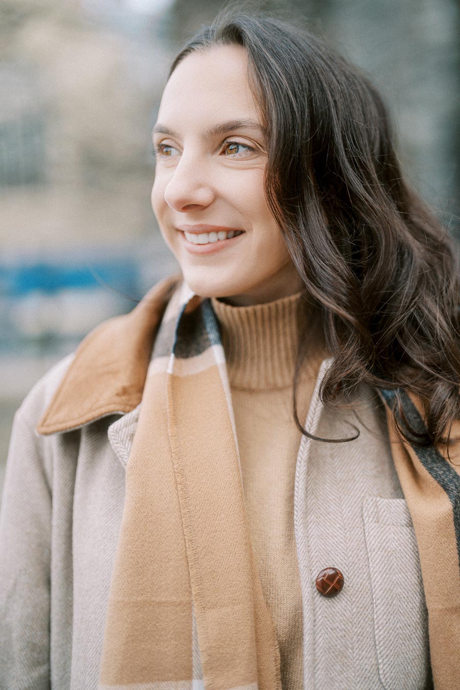 Smiling woman wearing a beige coat and scarf, enjoying a crisp outdoor setting.
