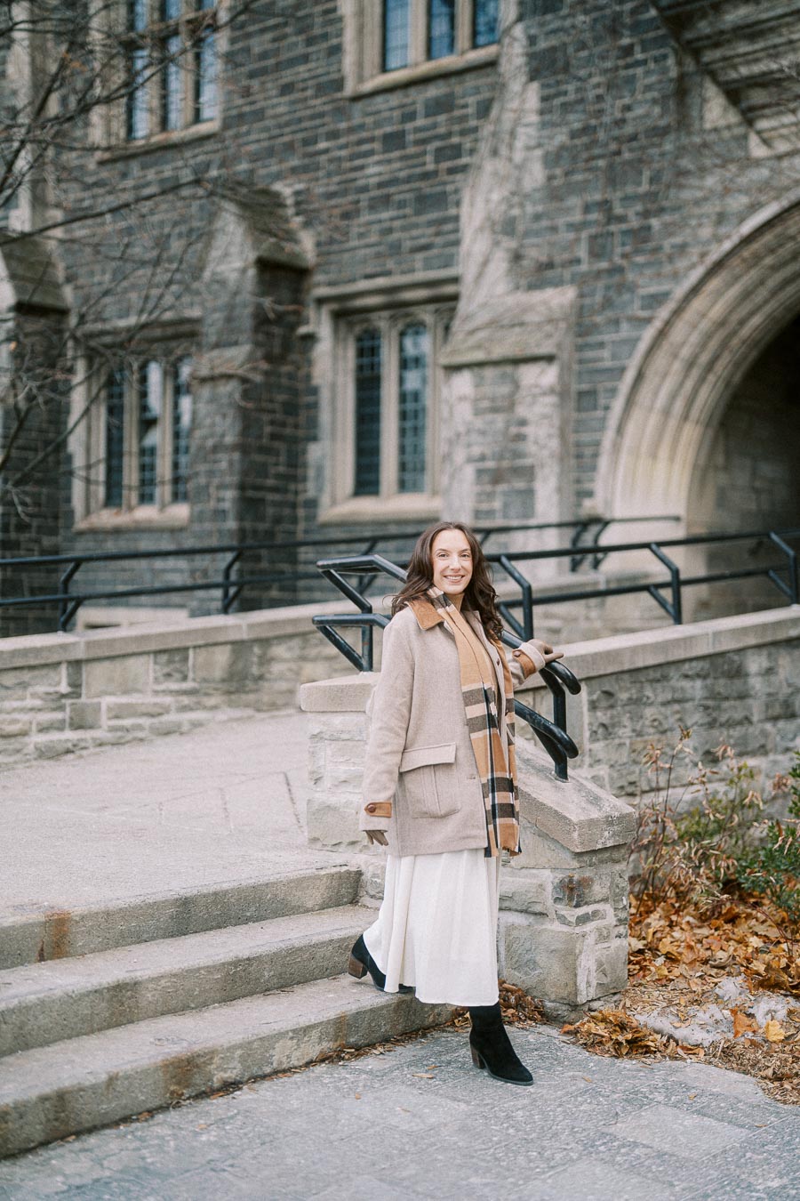 A woman wearing a light brown coat and scarf, smiling while walking down stone steps in front of a historic building with arched windows.