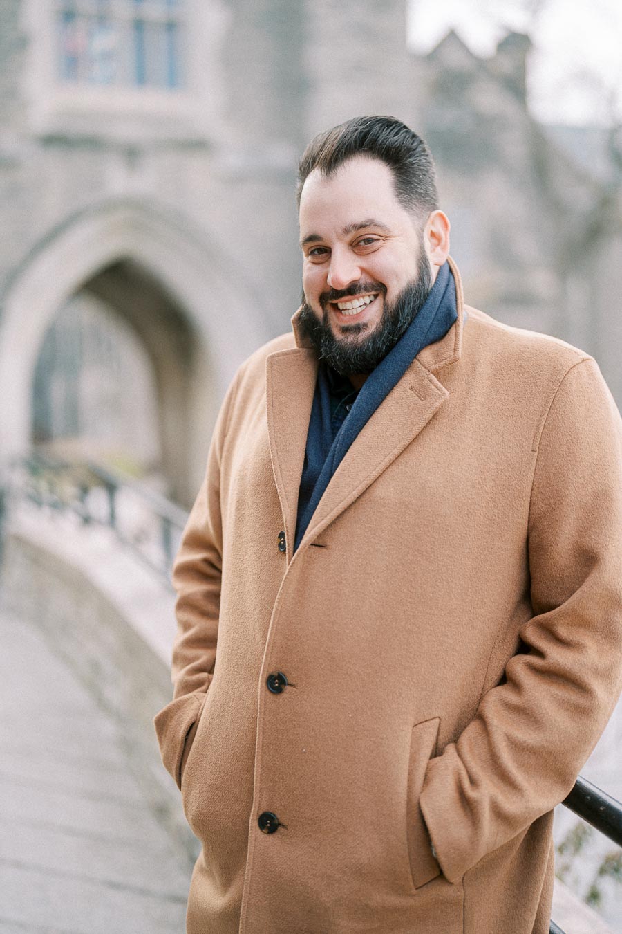 A person smiling while wearing a brown coat, standing outside near a stone building with an arch in the background.