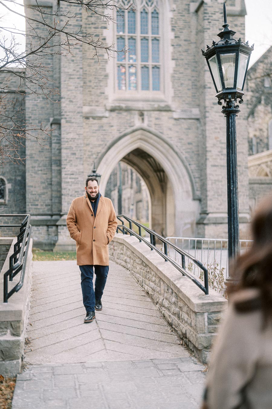A man in a brown coat walking on a stone pathway in front of a historic building with large arched windows and a black lamppost.