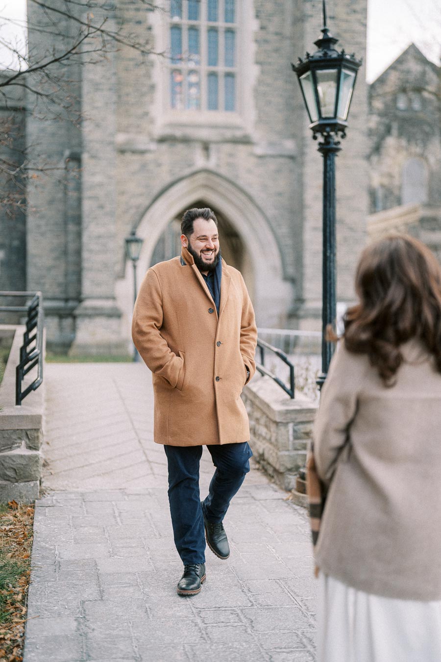 Man in a camel coat walking toward another person on a stone pathway outside a historic building with Gothic architecture and a black lamppost.