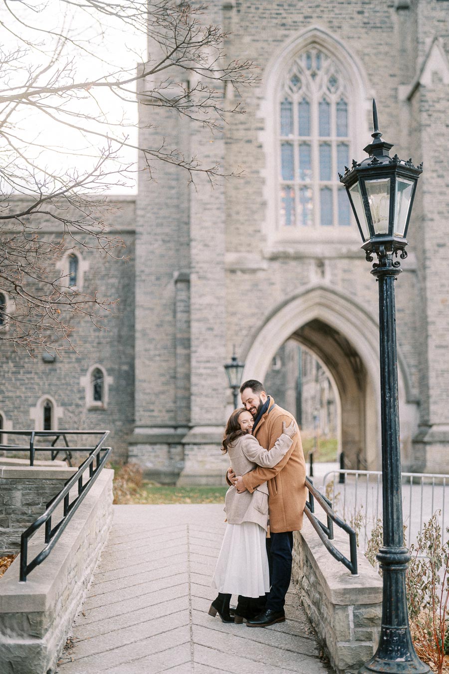 A couple embracing on a stone walkway in front of a historic building with arched windows, surrounded by winter trees and an ornate lamp post.